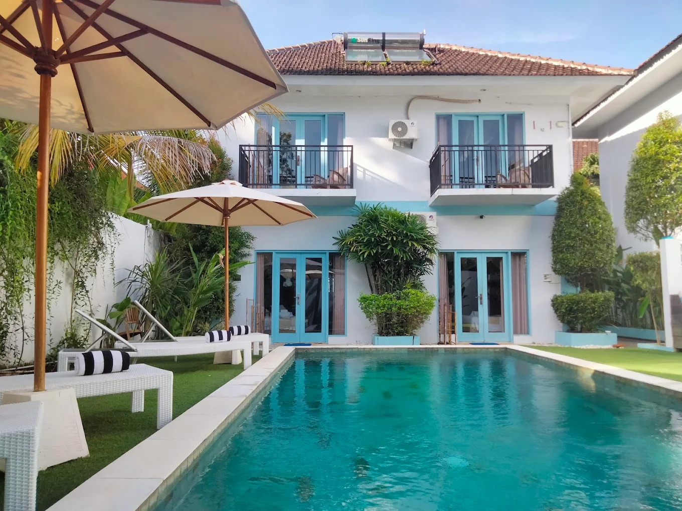 Two storey white villa with turquoise doors beside a turquoise pool and shaded loungers in Bali